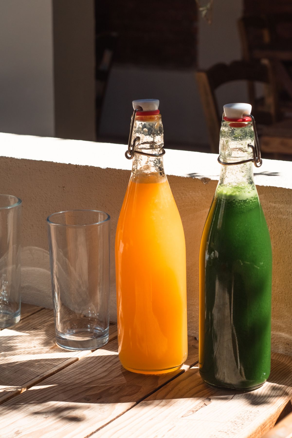 Two flip-top bottles of homemade kombucha in vibrant orange and green, sunlit on a wooden table