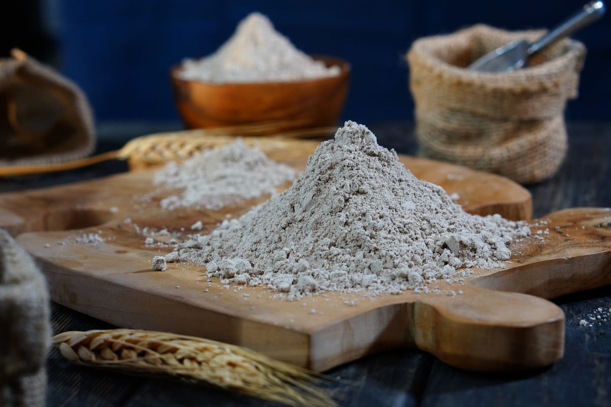 A mound of coarse wholemeal flour on a wooden board with wheat stalks and a grain sack beside it