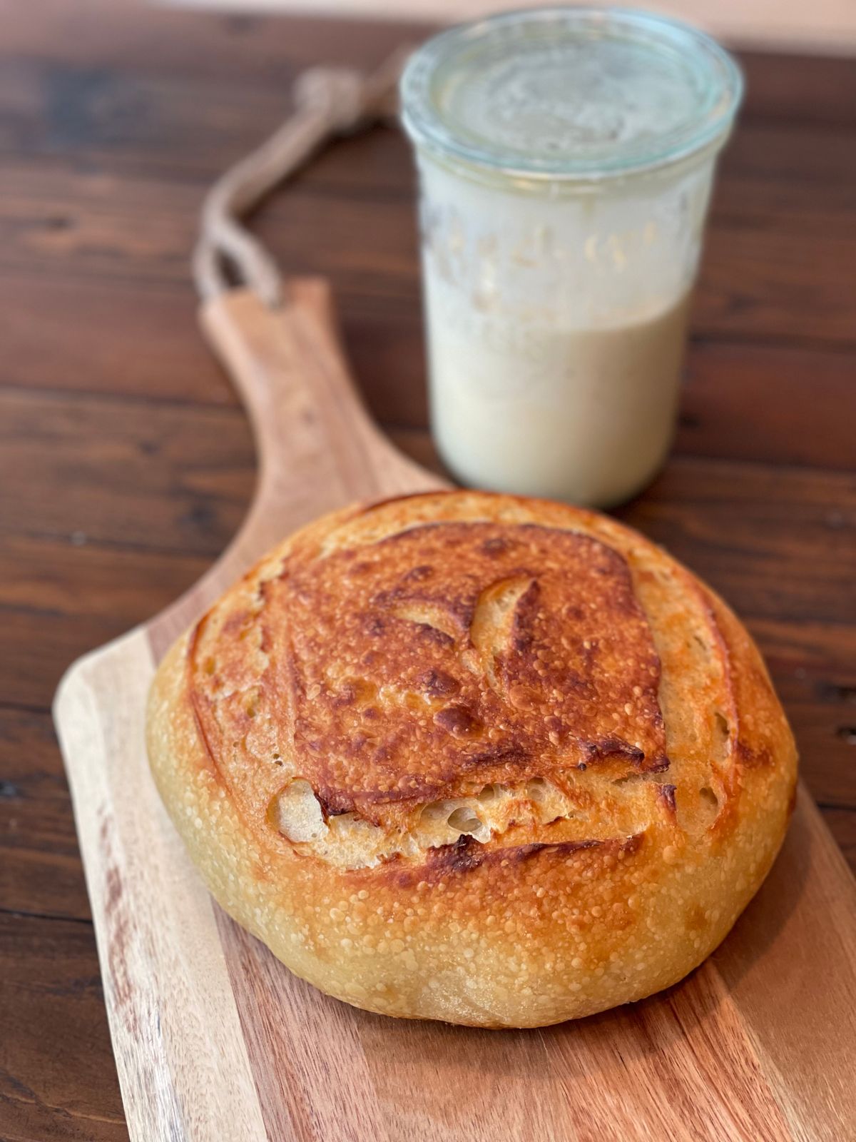 A round sourdough loaf on a wooden board beside a tall glass jar of starter