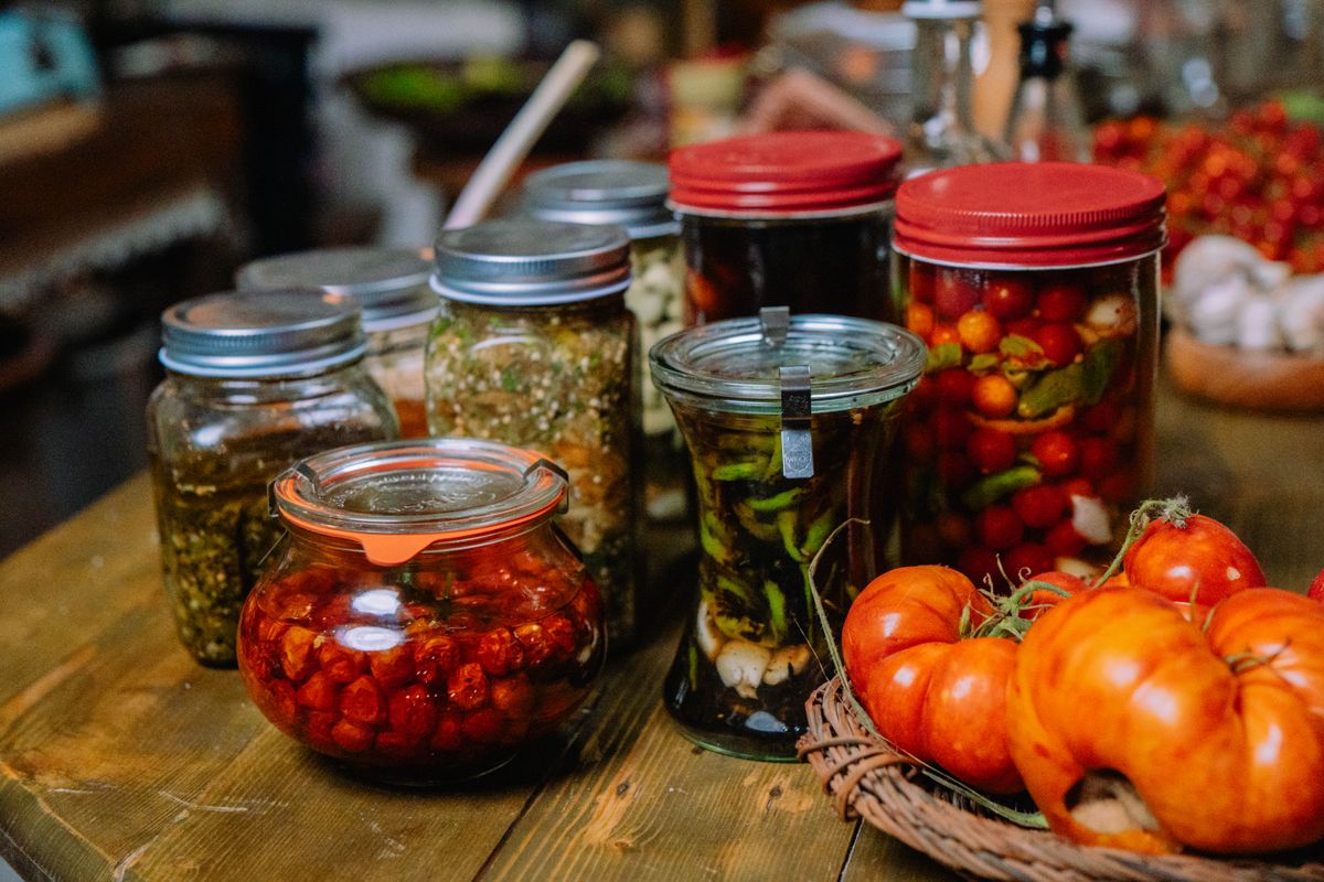 Assorted jars of preserved vegetables and fresh tomatoes spread across a rustic wooden table