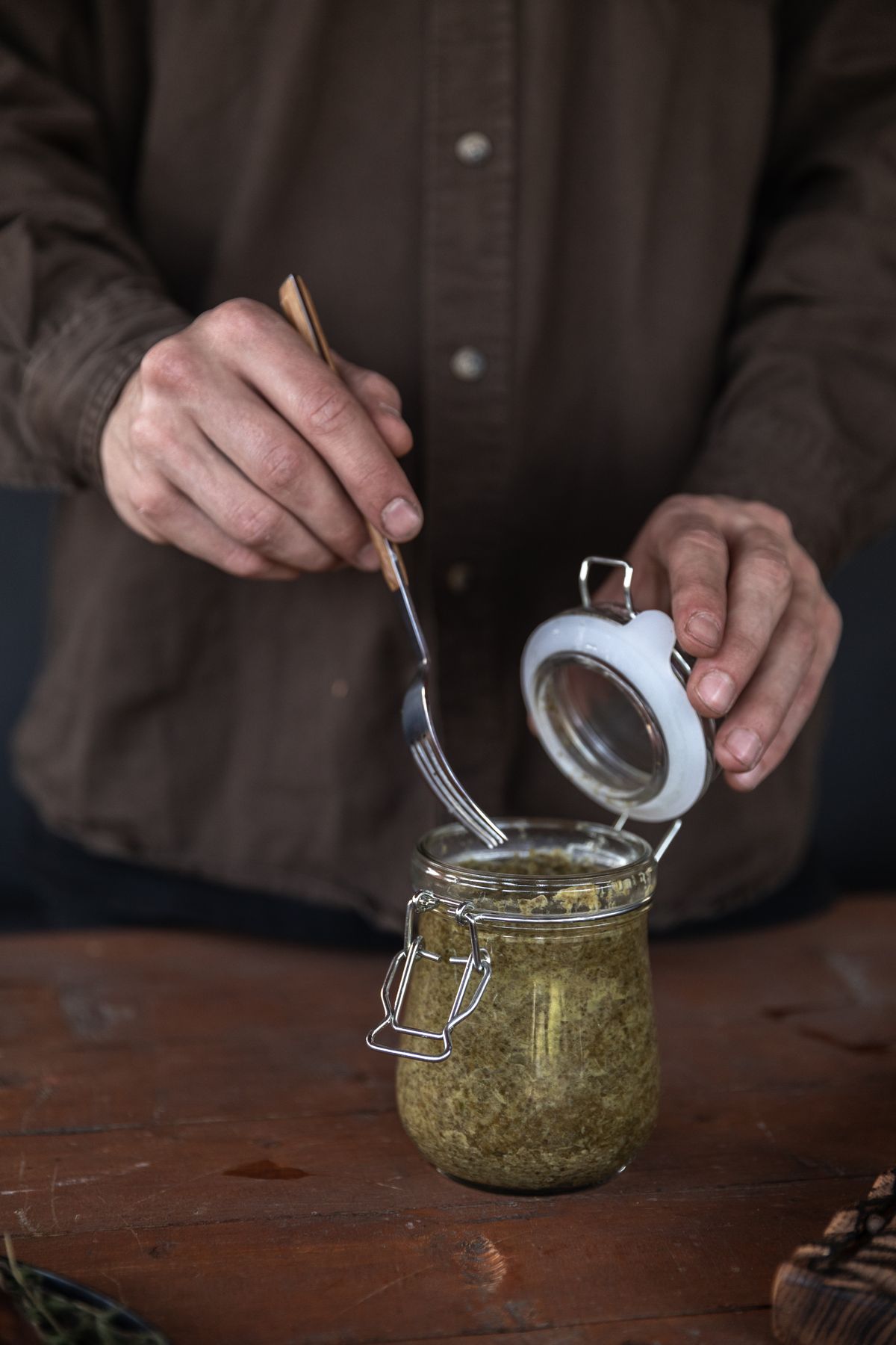Hands opening a clamp-lid jar of fermented vegetables with a fork at a wooden table