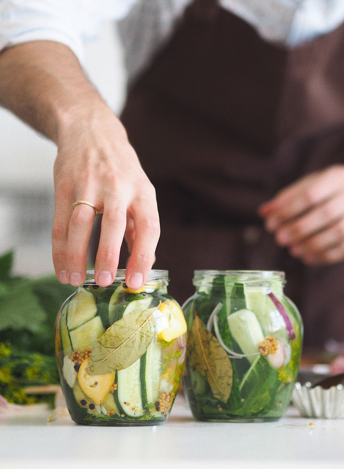 Hands reaching into a glass jar packed with sliced cucumbers, bay leaves and spices in brine