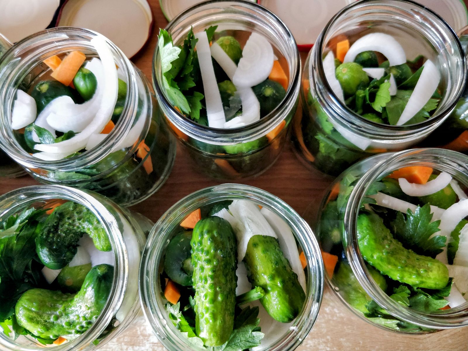 Six open glass jars packed with cucumbers, carrot, onion and parsley, seen from above on a wooden counter