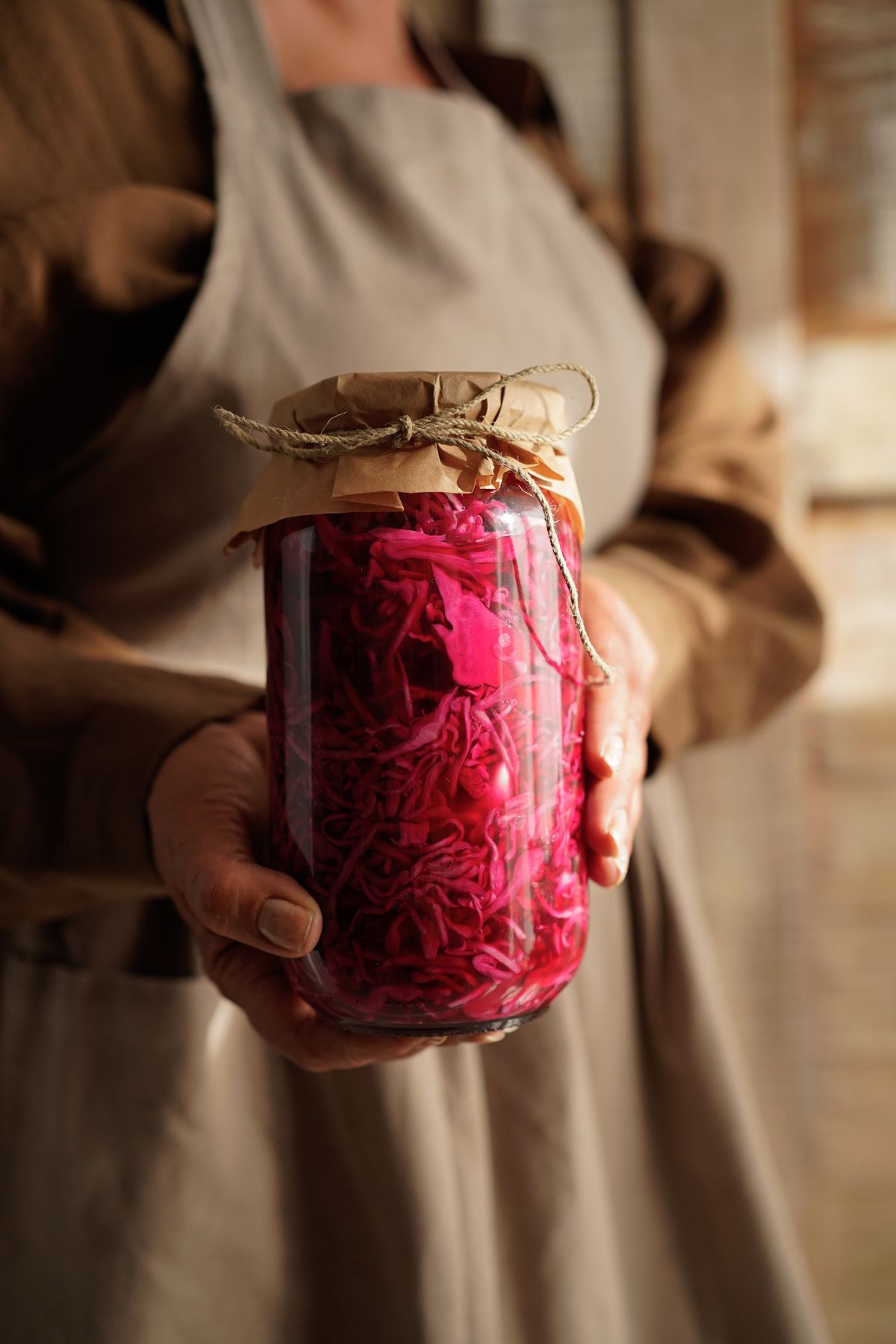 Two hands holding a tall jar of shredded pickled red cabbage with a paper cover tied over the lid