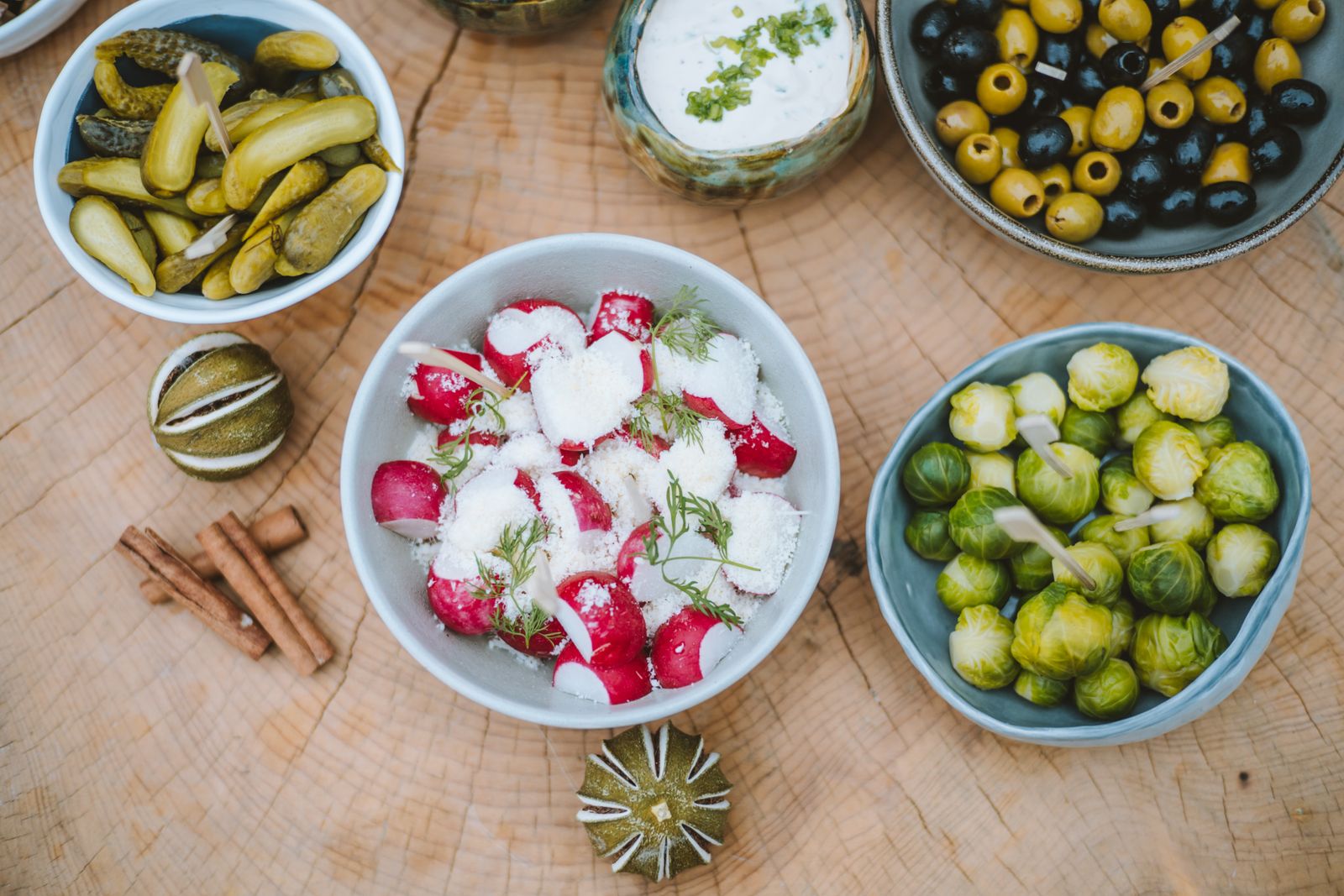 Bowls of gherkins, green olives, brussels sprouts and radish salad set out on a wooden table