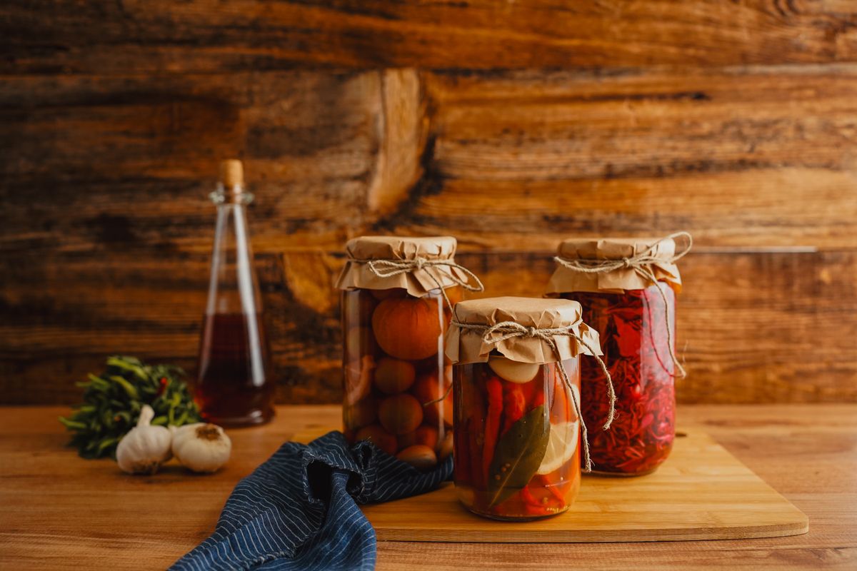 Three jars of preserved vegetables with brown paper covers on a wooden board, garlic and herbs alongside