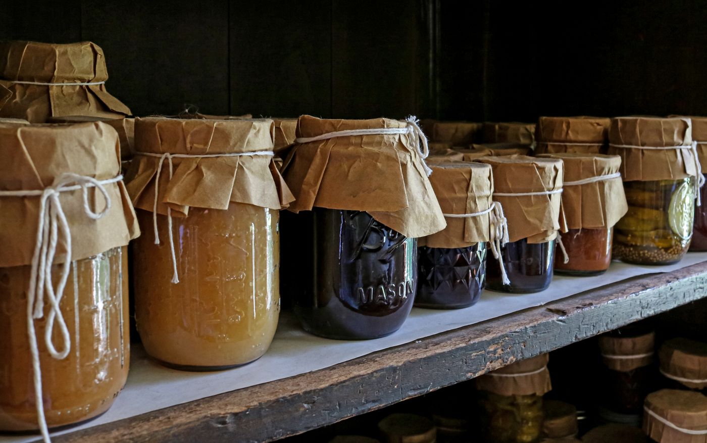 A wooden shelf lined with glass jars of preserved vegetables, brown paper tied over every lid