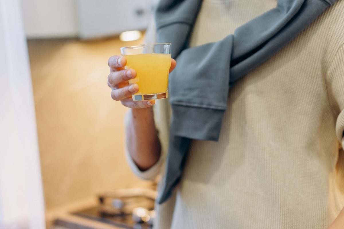 Hand holding a small glass of golden fermented drink in a sunlit kitchen