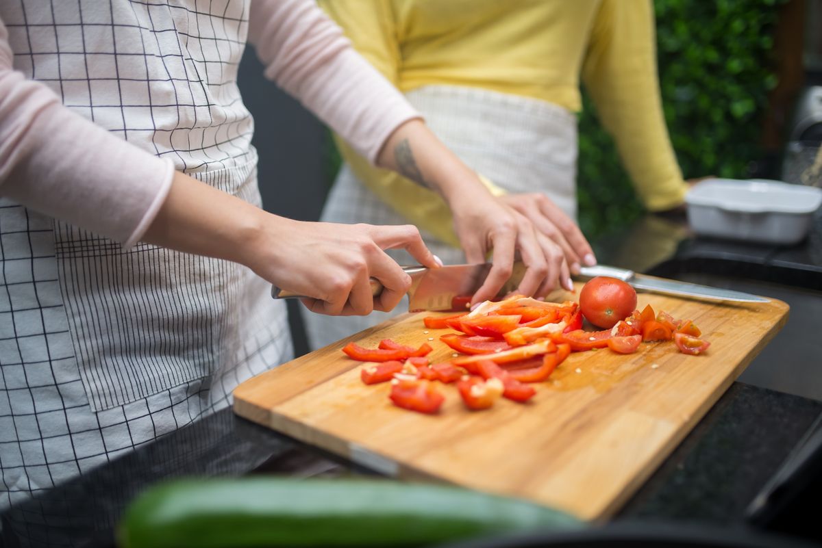 Hands chopping bright red tomatoes and green peppers on a wooden cutting board with a glass jar and kitchen scale nearby
