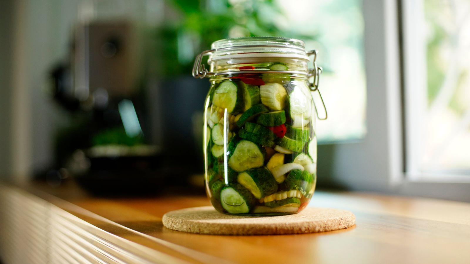 A tall clamp-lid jar of sliced cucumbers on a wooden kitchen counter, lit by a window