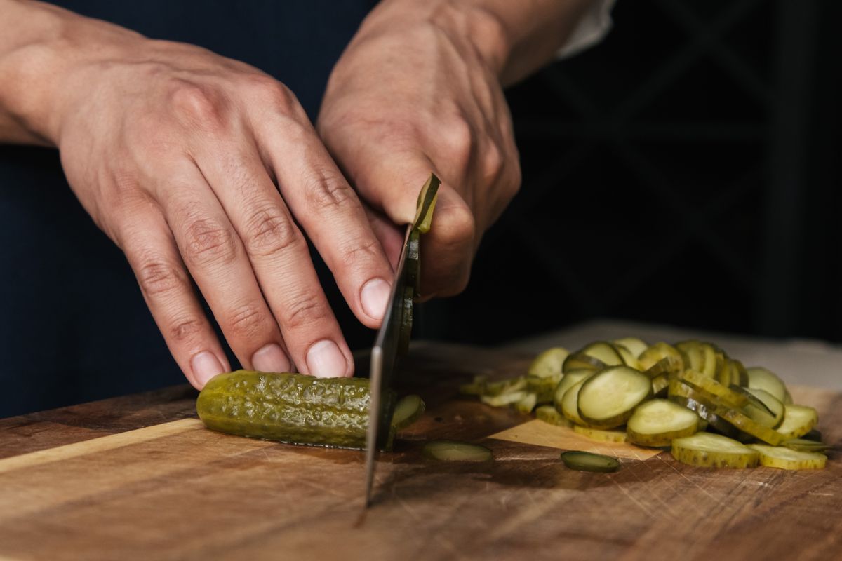 Hands slicing a brined cucumber into rounds on a wooden board